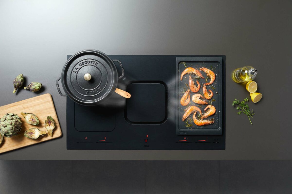 Zwarte ATAG inductiekookplaat met afzuiging, stoofpan en grillplaat; ingrediënten op donker werkblad zichtbaar.